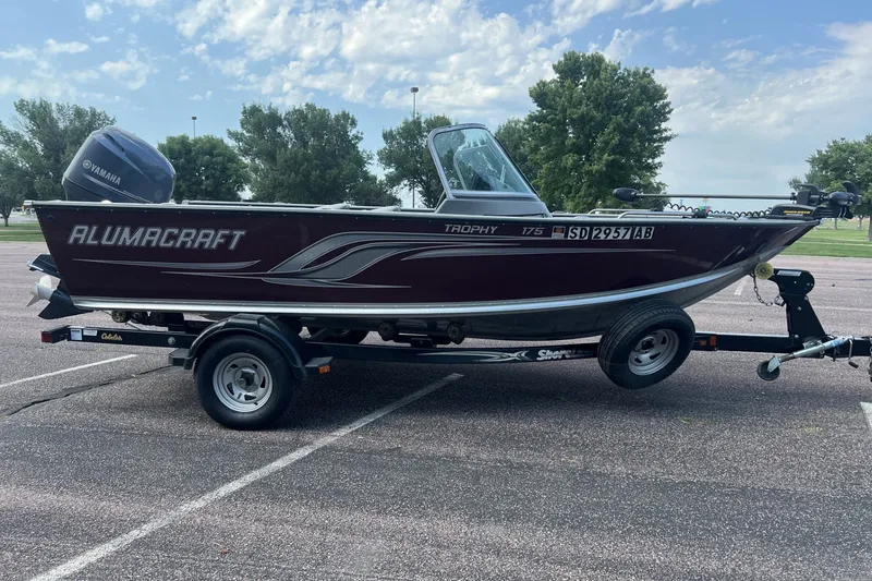Slide: The Image of 2012 Alumacraft Trophy 175 boat on trailer, parked in a lot under a cloudy sky. - 5