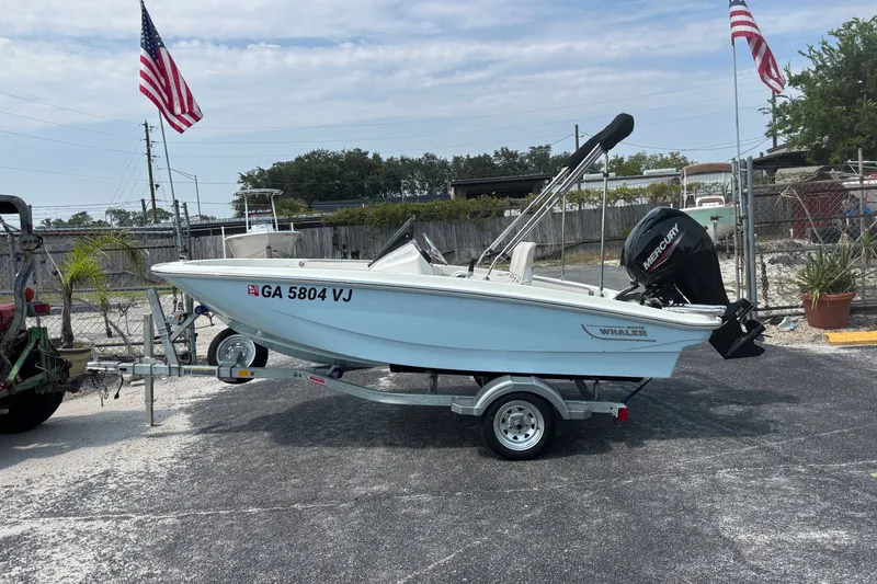 Slide: The Image of 2025 Boston Whaler 130 Super Sport boat on trailer, parked outdoors with American flags. - 8