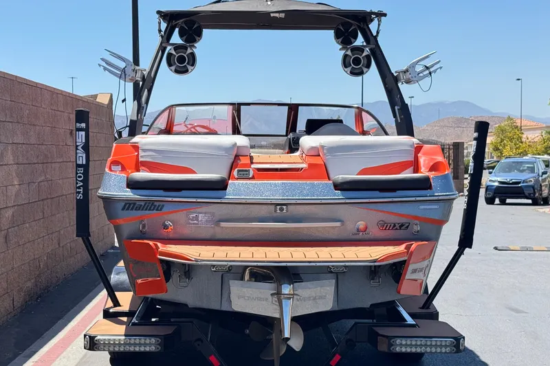 Slide: The Image of 2018 Malibu Wakesetter 22 MXZ boat, orange and silver, parked outdoors with speakers and wakeboard racks. - 4
