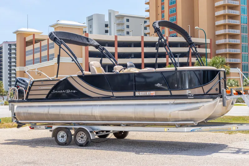 The Image of 2017 South Bay 523SL pontoon boat on trailer, parked near modern buildings. - 0