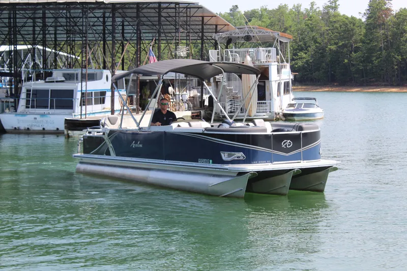 Slide: The Image of A 2024 Avalon LSZ RF 2585 pontoon boat on a calm lake. Boats are docked in the background. - 8