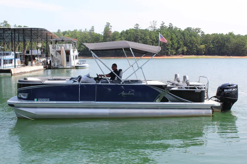 The Image of A 2024 Avalon LSZ RF 2585 pontoon boat on a calm lake with a sunshade and Suzuki outboard motor. - 1