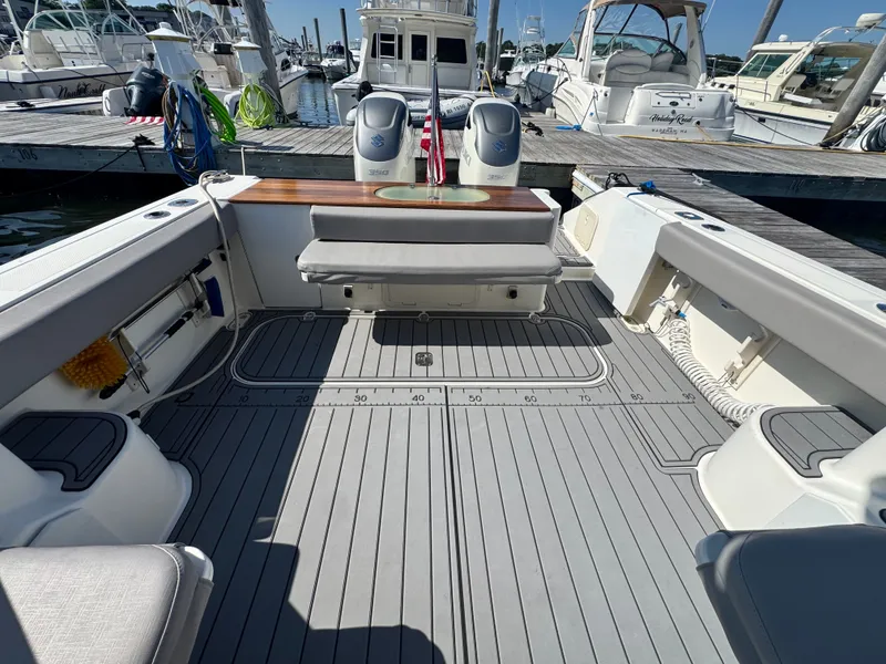 Slide: The Image of The image shows the cockpit of a 2006 Pursuit 3370 Offshore. It features a gray deck, a bench seat, and a work surface with a sink. Other boats are visible docked in the background. - 15