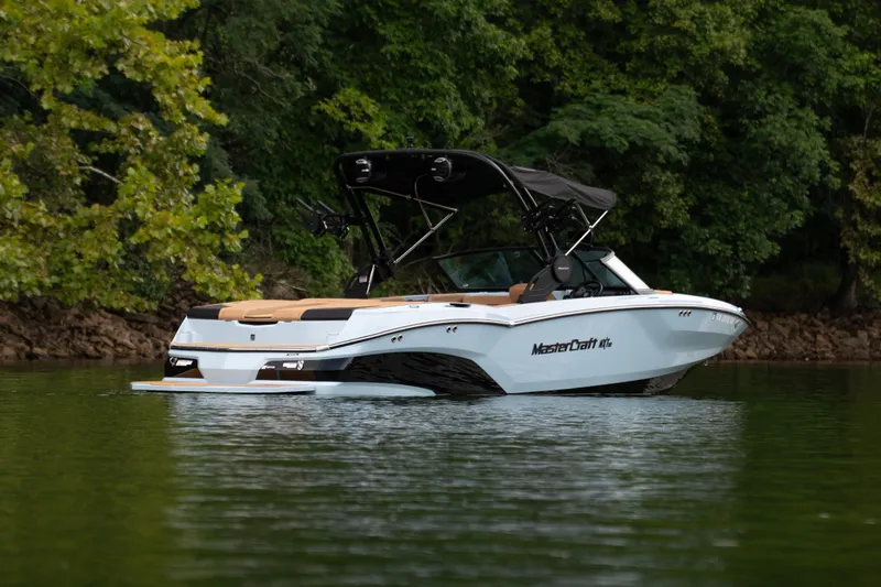 Slide: The Image of 2026 MasterCraft NXT23 boat on a serene lake with lush green trees. - 2