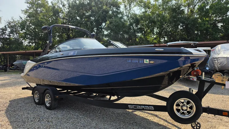 Slide: The Image of A blue 2019 Scarab 255 ID Wake Edition on a trailer, surrounded by trees. - 6