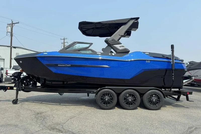 The Image of 2027 MasterCraft X23 boat in blue and black on a trailer, parked outdoors. - 1