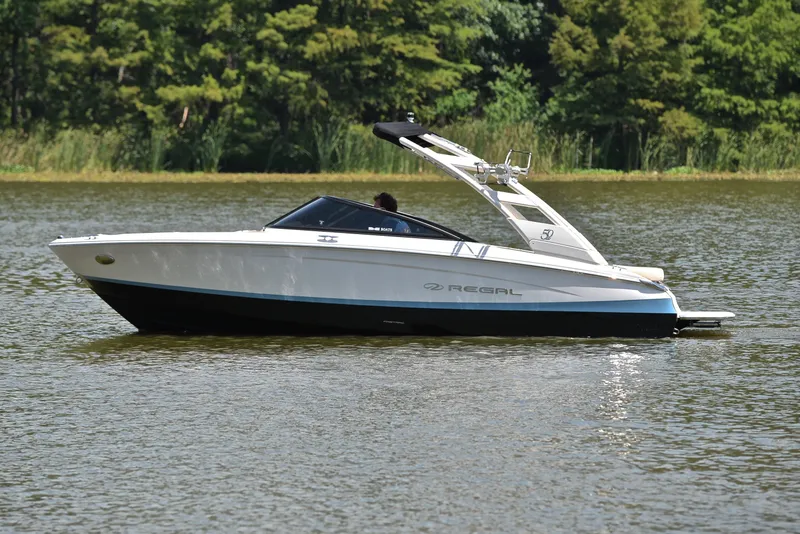 Slide: The Image of 2020 Regal LS2 Surf boat on a calm lake with lush greenery. - 10