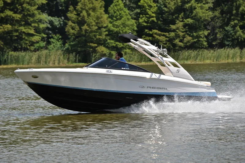 Slide: The Image of 2020 Regal LS2 Surf boat cruising on a lake with lush greenery in the background. - 2