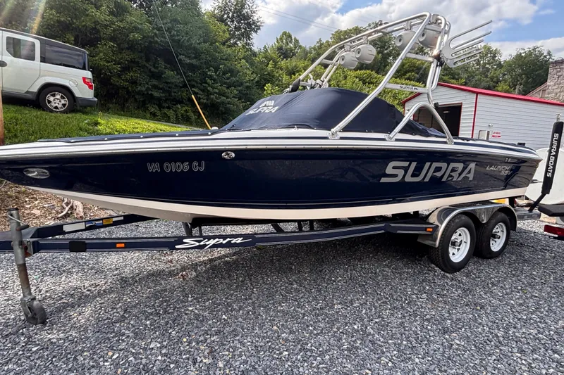 The Image of 2004 Supra Launch SSV boat on trailer, parked outdoors with trees and a building nearby. - 1
