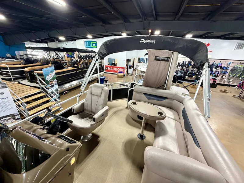 Slide: The Image of The image shows a 2011 Premier Sunsation 220 pontoon boat in a showroom. It features beige seating and a round table. - 6