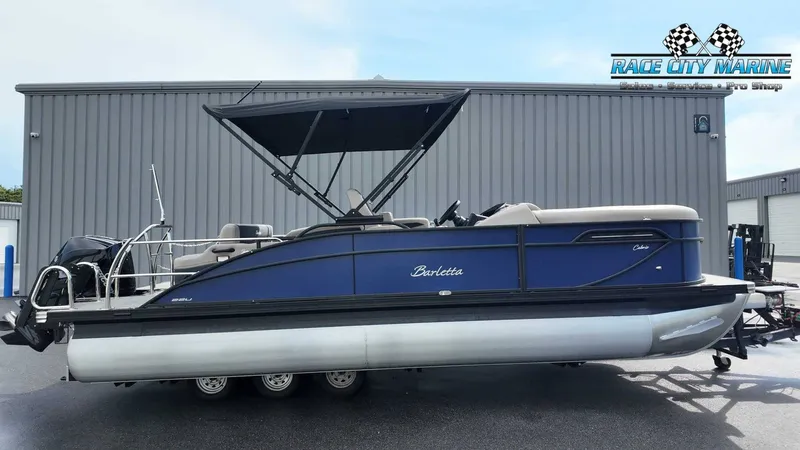 The Image of A 2027 Barletta C22U pontoon boat with a blue hull and black canopy, parked in front of a building. - 1