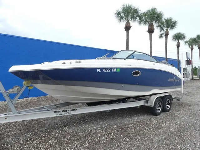 Slide: The Image of A blue and white 2023 NauticStar 243 DC on a trailer, with palm trees in the background. - 2