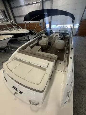 Slide: The Image of A 2013 Sea Ray SLX 230 in a warehouse. It features a black Bimini top and beige upholstery. - 10