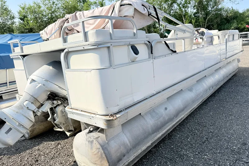 Slide: The Image of 1999 Crest 240 pontoon boat with outboard motor, parked on gravel, surrounded by trees. - 2