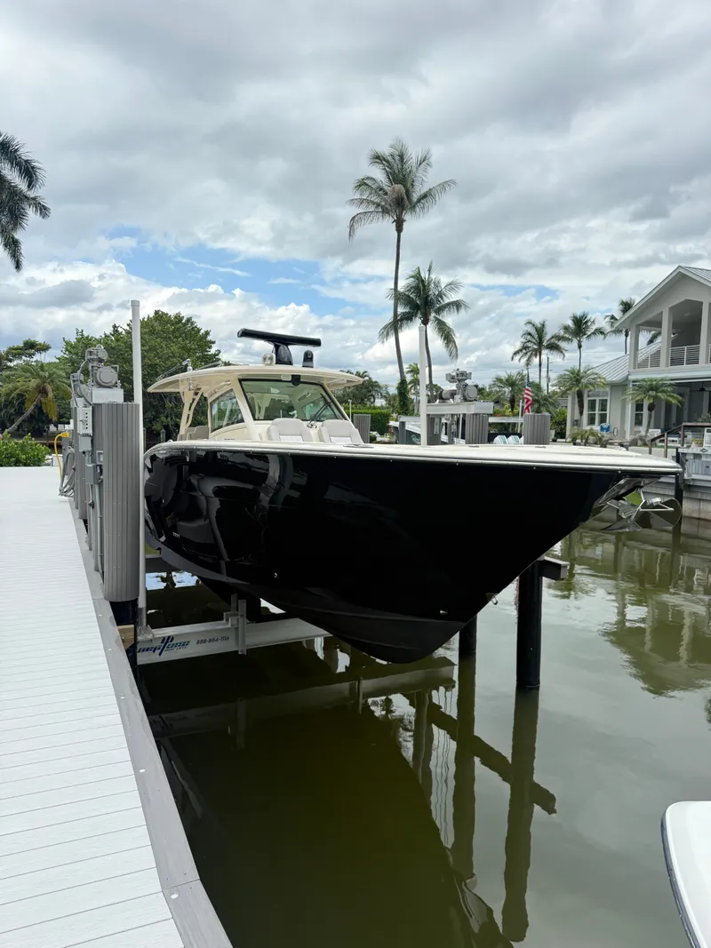 The Image of A 2019 Scout 420LXF boat is lifted at a dock. Palm trees and houses in the background. - 0