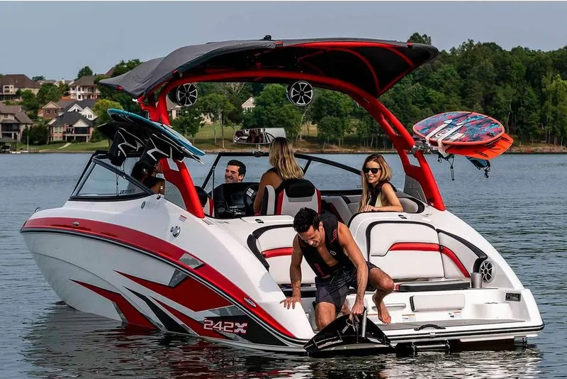 Slide: The Image of A 2020 Yamaha 242X boat on a lake. Four people enjoy the ride, with one man stepping onto the platform. A surfboard is attached to the boat. - 21