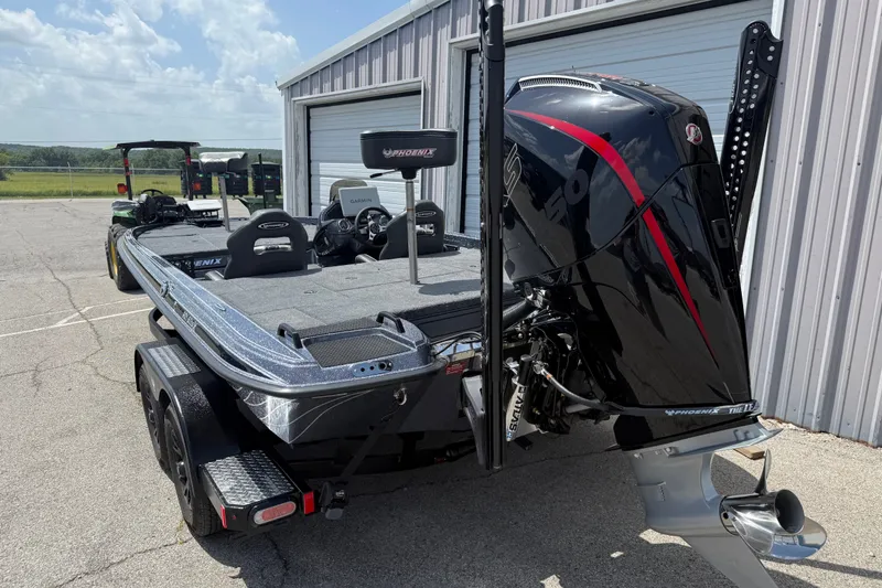 Slide: The Image of 2027 Phoenix 921 Elite X boat with powerful outboard motor, parked near a metal building. - 2