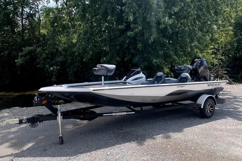 The Image of A 2023 Vexus AVX189C boat on a trailer, surrounded by trees and gravel ground. - 1