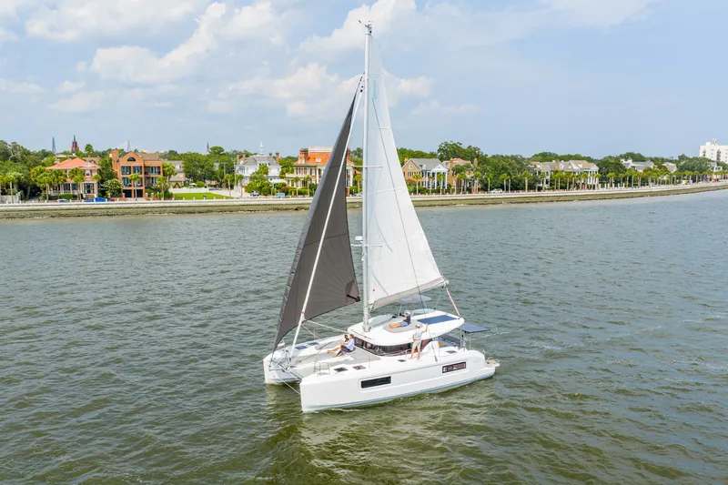 Slide: The Image of 2026 Lagoon 38 catamaran sailing near a scenic coastal town under a partly cloudy sky. - 100