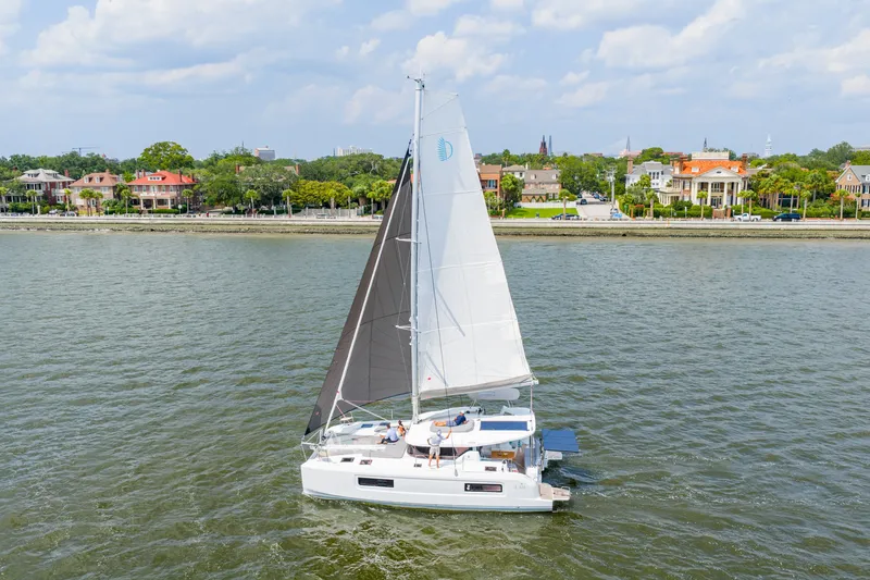 Slide: The Image of 2026 Lagoon 38 catamaran sailing near a scenic coastal town under a partly cloudy sky. - 99
