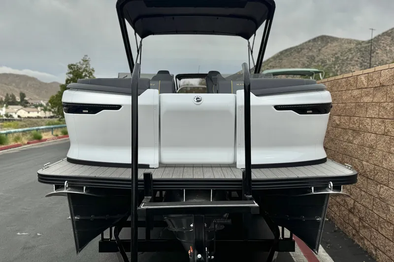 Slide: The Image of 2025 Manitou EXPLORE 24 pontoon boat, rear view, parked on a road with scenic background. - 2