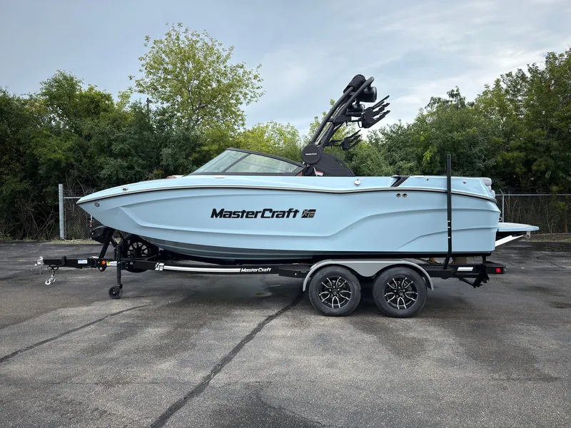 The Image of A 2027 MasterCraft XT20 boat on a trailer, surrounded by trees. - 0