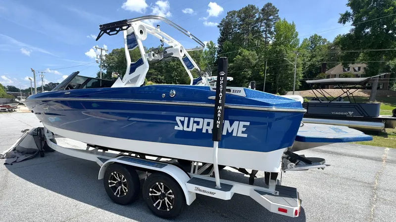 Slide: The Image of A blue 2022 Supreme S220 boat on a trailer. It features a white tower and is parked on a lot. - 4