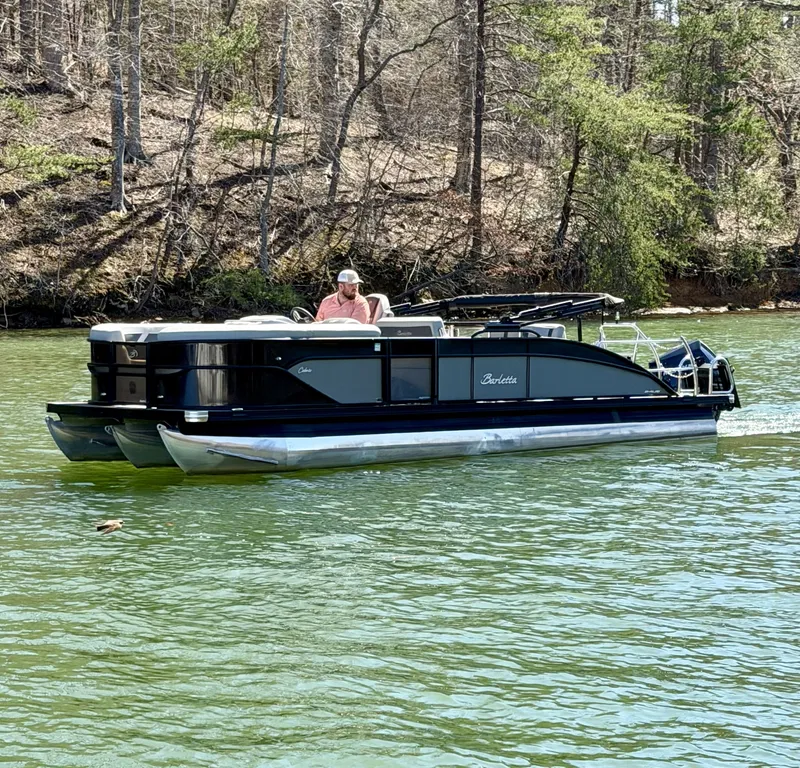 The Image of A Barletta Cabrio 24 UE pontoon boat cruising on a calm lake, surrounded by trees. - 1