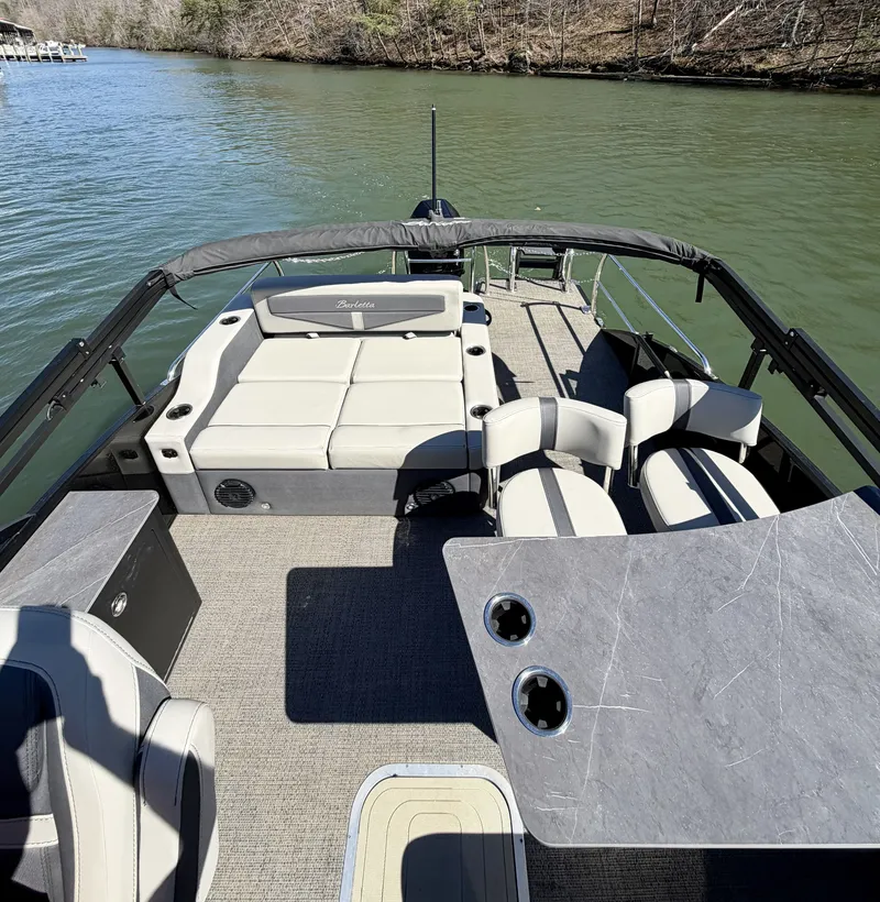 Slide: The Image of The image shows a 2024 Barletta Cabrio 24 UE pontoon boat on calm water. It features spacious seating with comfortable cushions and a table with cup holders. The surroundings are tree-lined. - 8