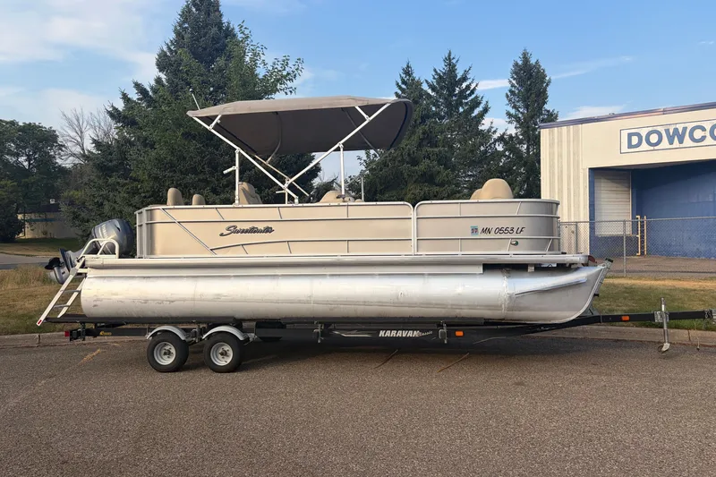 The Image of 2015 Sweetwater 2286 pontoon boat on trailer, parked outdoors near trees and a building. - 0