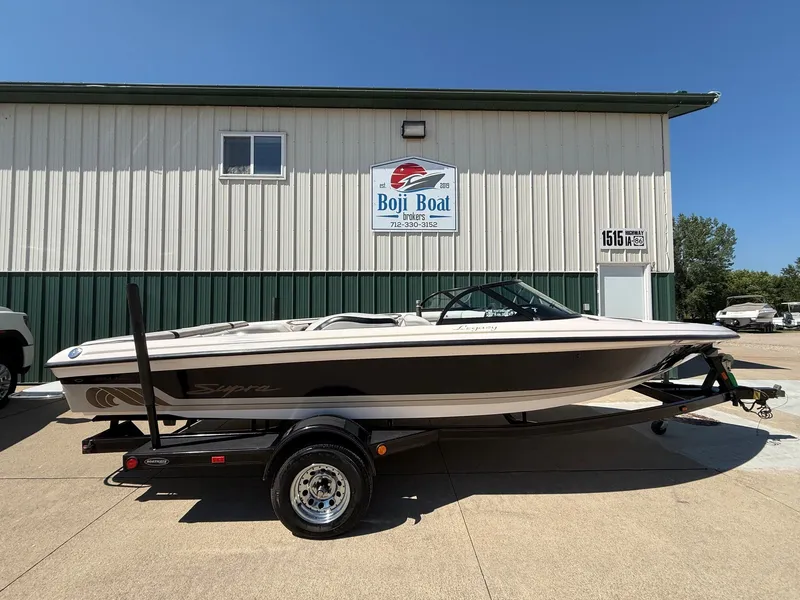 The Image of A white and black 1999 Supra Legacy boat on a trailer in front of a building. - 0