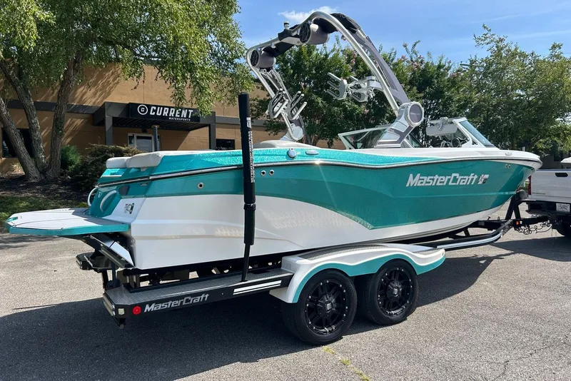 Slide: The Image of 2017 MasterCraft XT23 boat on trailer, teal and white, parked outdoors near trees. - 2