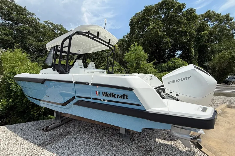 Slide: The Image of A 2026 Wellcraft 28 T-Top boat on a gravel bed, surrounded by trees. - 1