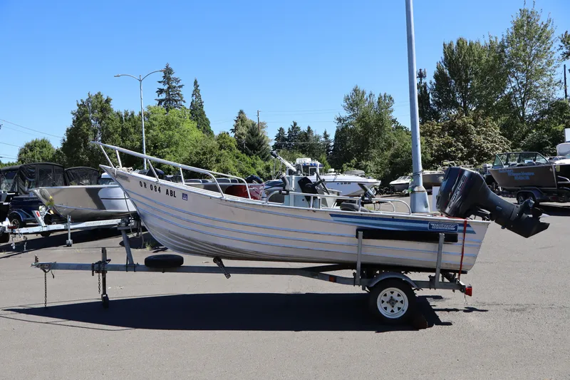 Slide: The Image of 1985 Klamath 15 boat on trailer in outdoor lot, surrounded by trees and other boats. - 2