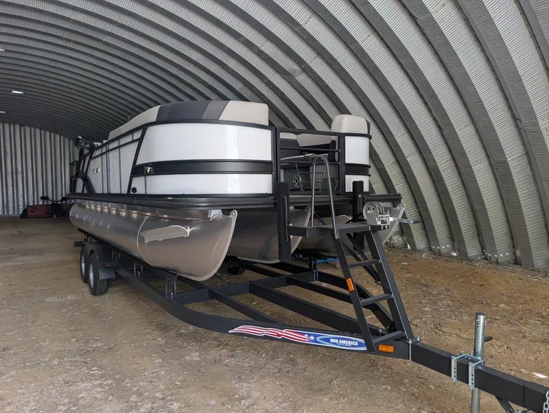 Slide: The Image of A 2026 Sylvan L3 LZ DH pontoon boat on a trailer inside an arched warehouse. - 0