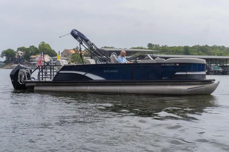 The Image of 2024 Sylvan L5 DLZ pontoon boat cruising on a lake with a driver onboard. - 1