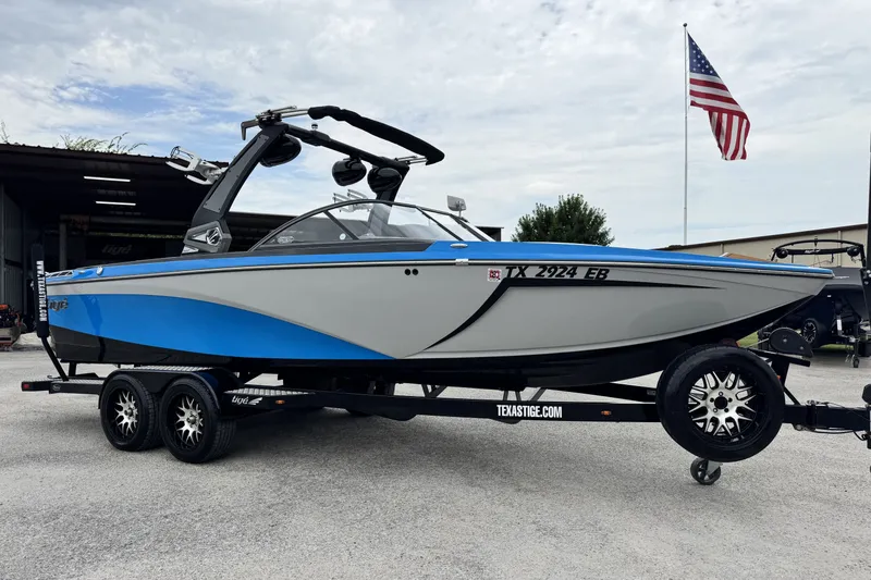Slide: The Image of 2017 Tigé R23 boat on trailer, blue and gray design, parked near American flag. - 1