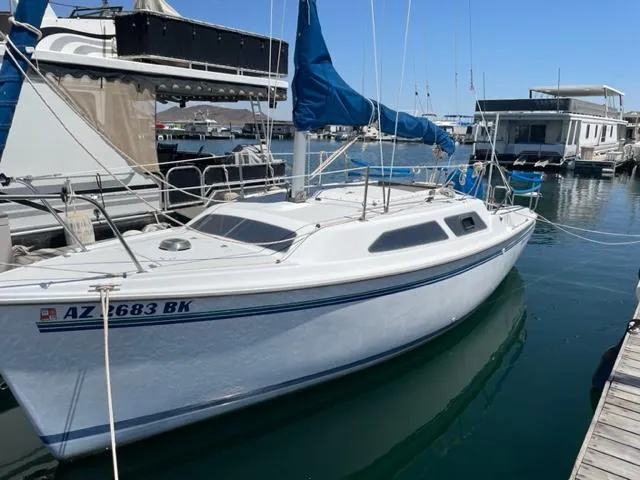 The Image of A white 1998 Catalina 250 sailboat is docked in a marina, featuring a blue sail. - 0