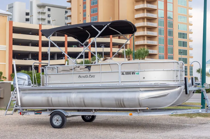 The Image of 2016 South Bay 5220CR pontoon boat on trailer, parked near modern buildings. - 0