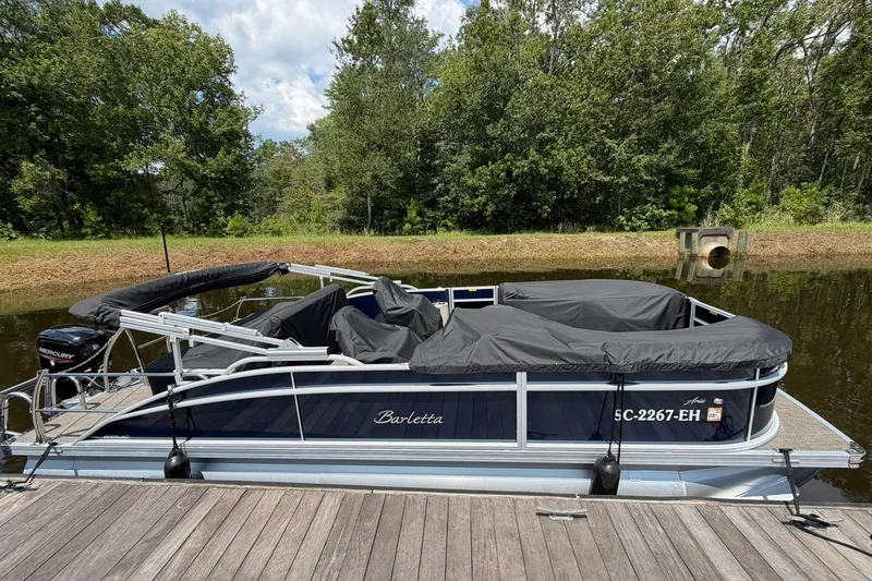 The Image of 2024 Barletta A22UC pontoon boat docked by a serene lake with lush greenery. - 0