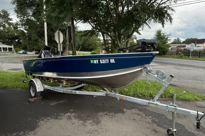 The Image of 2024 Lund 1600 Fury Tiller boat on trailer, parked roadside with trees in background. - 0