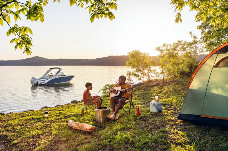 Slide: The Image of A 2027 Sea Ray SDX 230 Outboard is moored by a lakeside. Two people sit on the shore, one playing guitar while the other listens. A tent is nearby, and the sun is setting. - 19