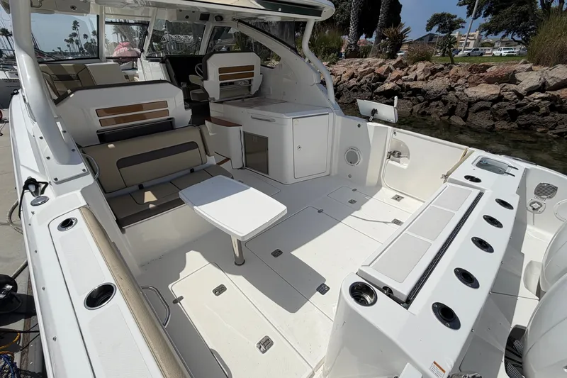 Slide: The Image of 2019 Pursuit DC 365 Dual Console boat interior with seating and table, docked by rocky shoreline. - 15