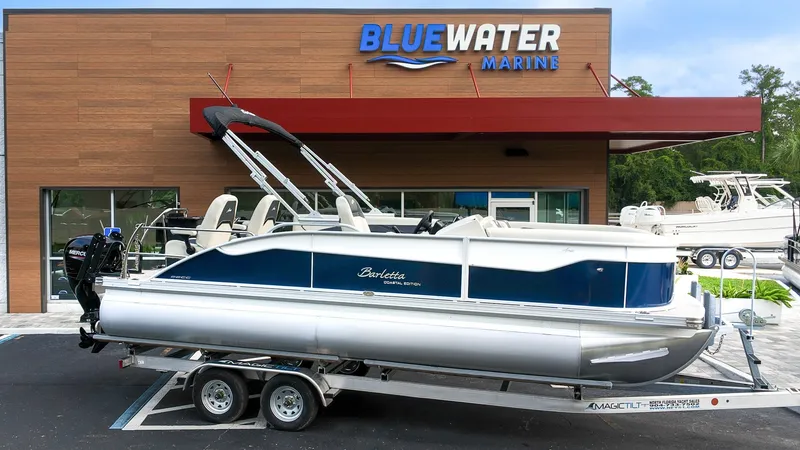 The Image of A 2027 Barletta Aria 22CC pontoon boat is on a trailer in front of a Bluewater Marine building. - 1
