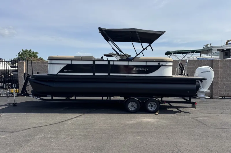 The Image of 2027 Godfrey Xperience 23' Split Bench pontoon boat on trailer, parked outdoors. - 0