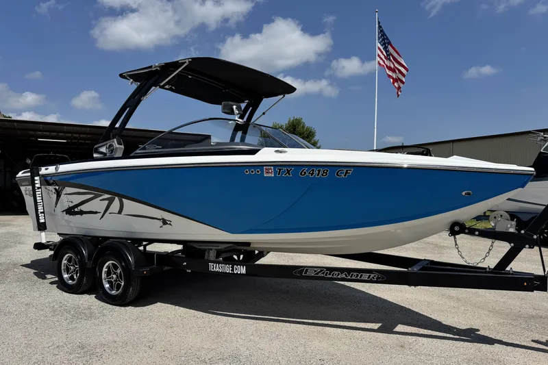 Slide: The Image of 2014 Tigé Z1 boat on trailer, blue and white design, parked outdoors with American flag. - 2
