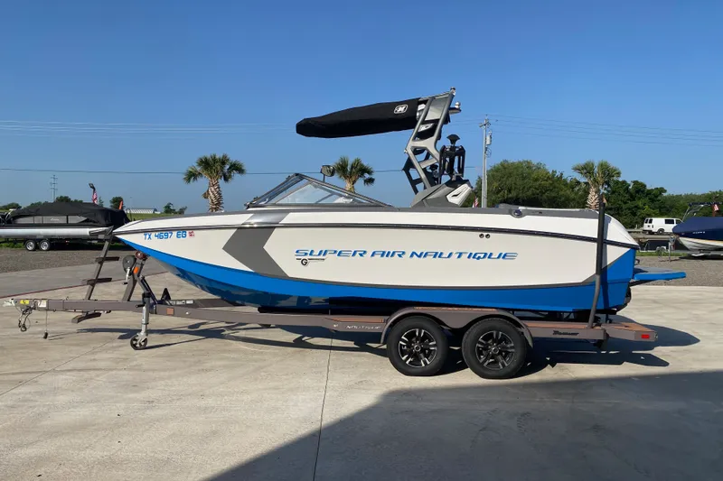 The Image of 2018 Nautique Super Air Nautique G21 boat on trailer, blue and white design, sunny day. - 12