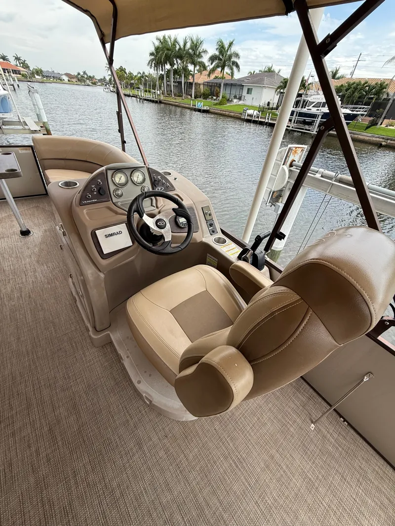 Slide: The Image of The image shows the helm of a 2018 Veranda VVF25F2. It features a beige carpeted floor and a comfortable, cushioned captain's chair. In the background, there's a calm waterway with palm trees and houses. - 2