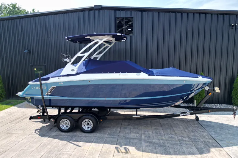 Slide: The Image of 2022 Cobalt R4 Surf boat on trailer, blue cover, parked by black building. - 27