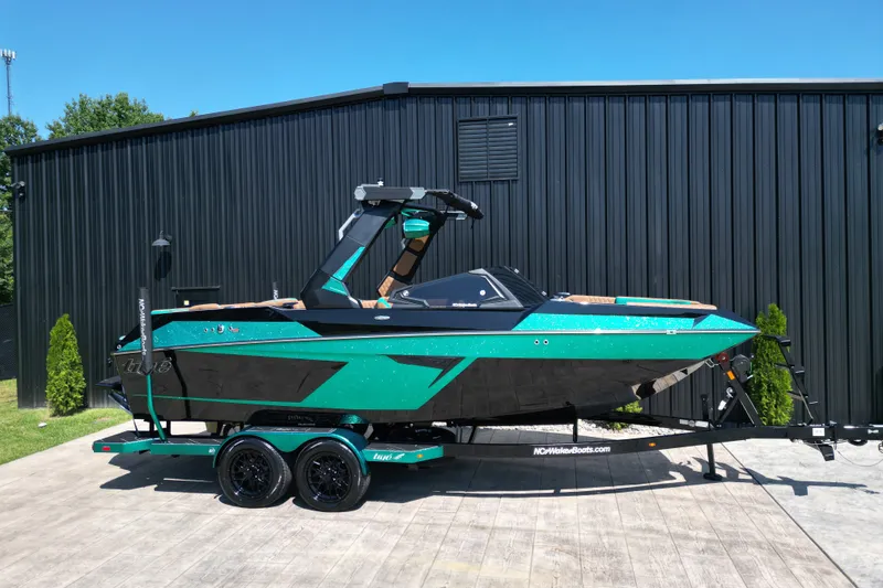 Slide: The Image of 2027 Tigé 22RZX boat in teal and black, parked on a trailer beside a black building. - 22